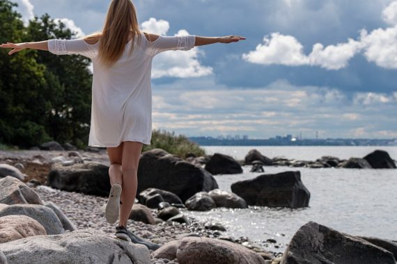 A blonde girl in a white summer dress stands on seaside rocks at the Viimsi Open Air Museum with her arms outstretched like wings, embracing the open air and the calm beauty of the Baltic Sea.